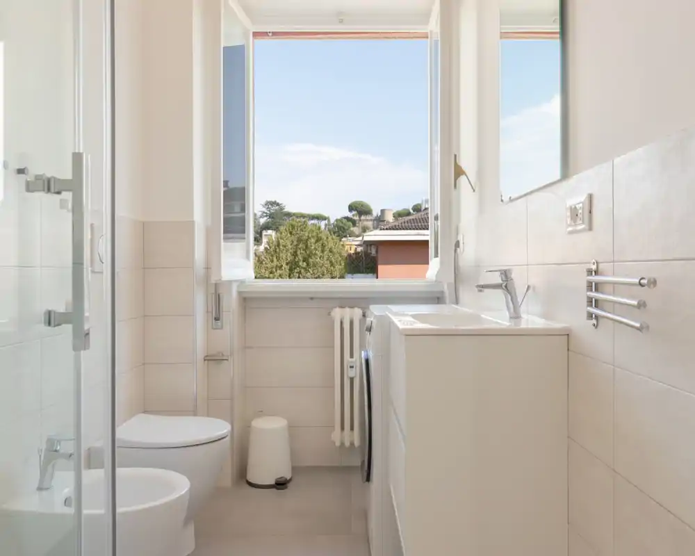 Salle de bains moderne et lumineuse avec douche vitrée, toilettes et bidet suspendus, meuble vasque et une grande fenêtre ouverte laissant entrer la lumière naturelle et offrant une vue.