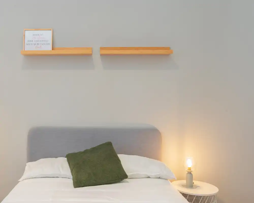 Minimal, softly lit bedroom with a grey headboard, white bedding and a green accent cushion; a small bedside table with a warm lamp creates a cozy atmosphere.