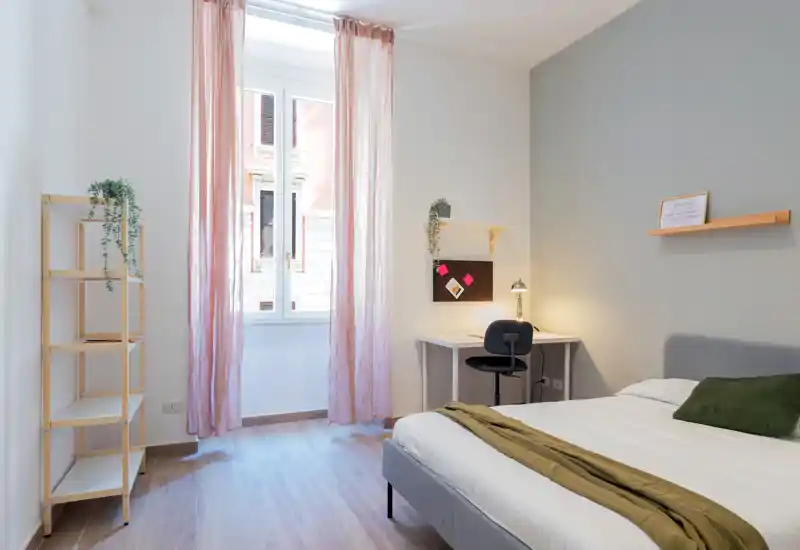 Camera da letto luminosa e minimal con un comodo letto matrimoniale, tessili morbidi e una scrivania vicino alla finestra — luce naturale e tonalità pastello per un ambiente moderno e rilassante.
