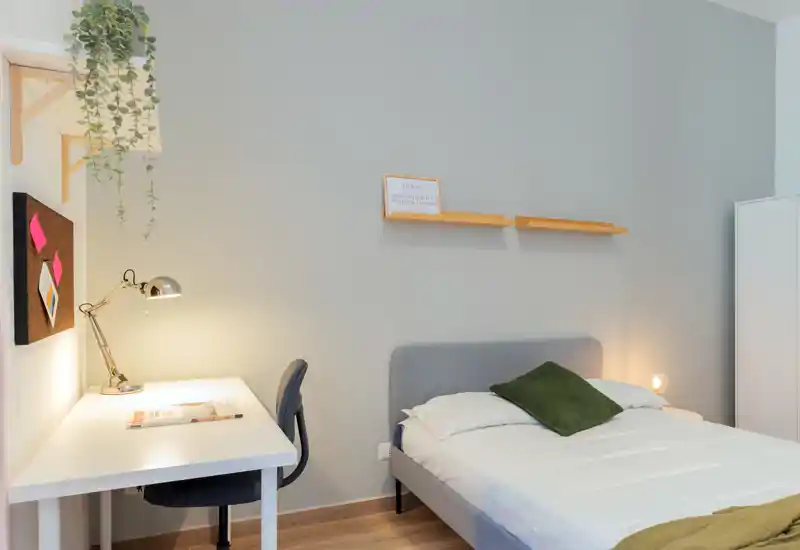 Camera da letto moderna e luminosa con letto matrimoniale ben rifatto, lampada da comodino e una scrivania per studio — toni neutri e una pianta sospesa rendono l'ambiente accogliente.
