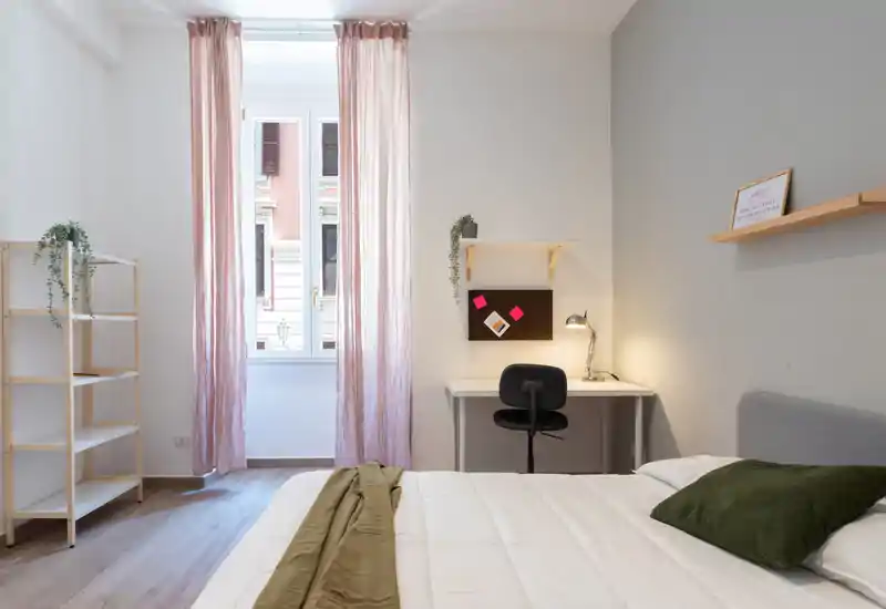 Camera da letto luminosa e minimale con un comodo letto in primo piano, una piccola scrivania vicino alla finestra, tende rosa e mensole semplici — ideale per un soggiorno rilassante o un breve affitto lavorativo.