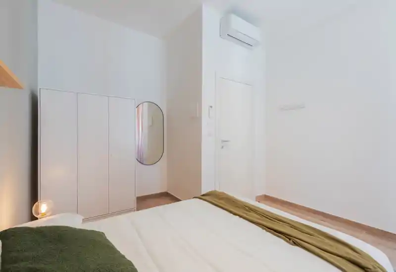 Camera matrimoniale luminosa e minimalista con letto ben rifatto, armadio, specchio ovale e lampada da comodino, perfetta per soggiorni rilassanti.