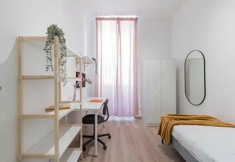 Camera da letto luminosa e minimal con letto singolo, armadio e scrivania vicino alla finestra con tende rosa leggere — ideale per uno studente o una persona sola.
