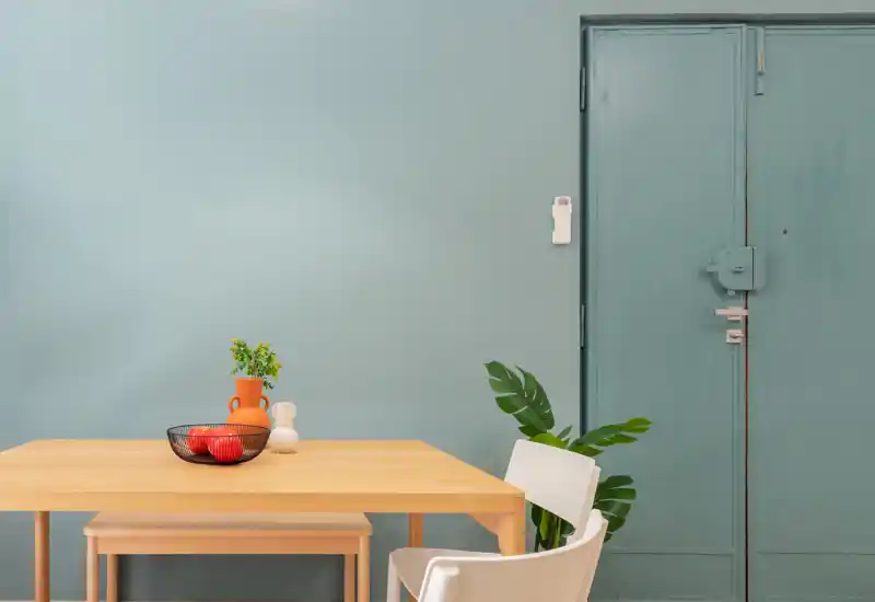 Coin repas minimaliste avec table en bois clair, banc et chaise, décoré d'un bol de fruits, d'un vase et d'une plante verte sur un mur bleu doux.