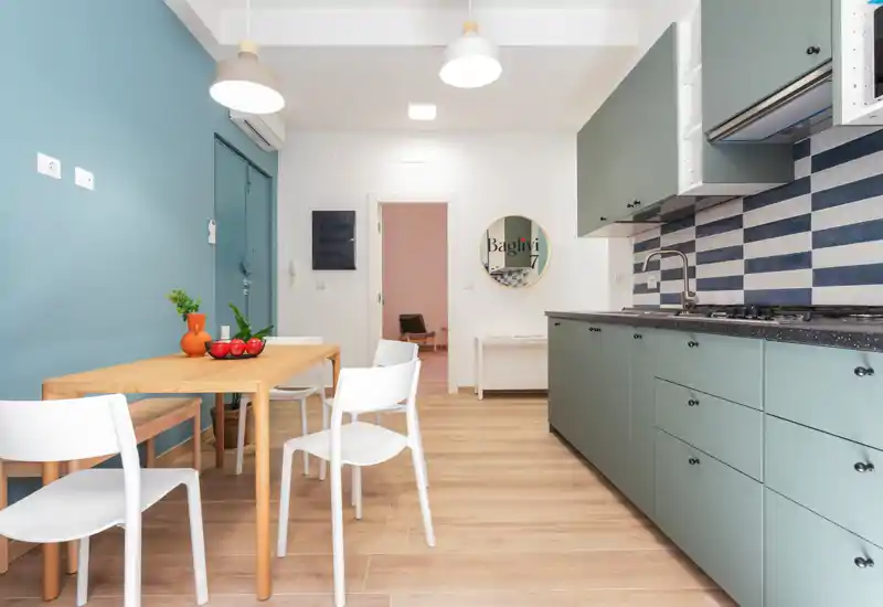 Cuisine moderne et lumineuse avec meubles vert menthe, crédence rayée et table en bois avec chaises blanches — un espace soigné, parfait pour les locations de courte durée.