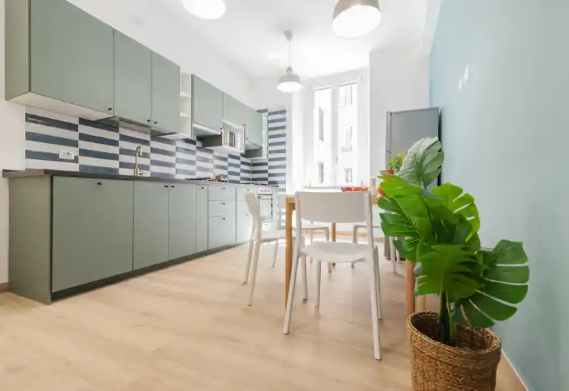 Cucina moderna e luminosa con pensili verde salvia, paraschizzi a righe e un piccolo tavolo da pranzo — ben illuminata e arredata con gusto, perfetta per una casa urbana compatta.
