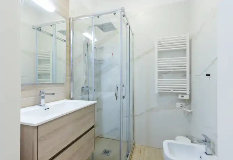 Salle de bains moderne et compacte avec cabine de douche en verre, radiateur sèche-serviettes mural, meuble vasque avec grand miroir — carrelage effet marbre et tons bois clairs pour une atmosphère contemporaine et fraîche.