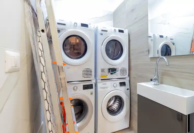 Petite buanderie/atelier avec lave-linge et sèche-linge empilés, un petit lavabo et une échelle sur le côté — fonctionnel et bien éclairé.