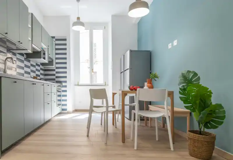 Cucina moderna e luminosa con pensili verde menta, paraschizzi a righe, tavolo da pranzo e sedie vicino a una finestra ampia — ambiente pulito e accogliente con luce naturale.