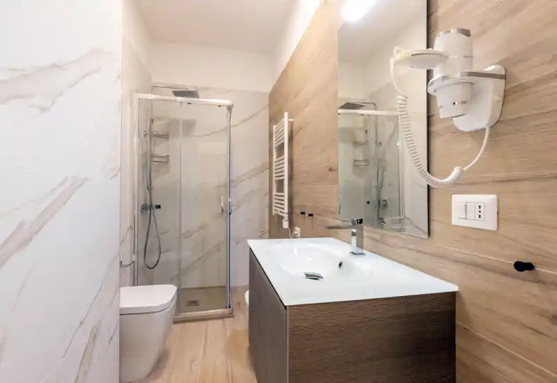 Salle de bains moderne avec cabine de douche en verre, sèche-cheveux mural, meuble vasque et toilettes. Carrelage effet marbre et finitions bois claires pour une ambiance contemporaine et chaleureuse.