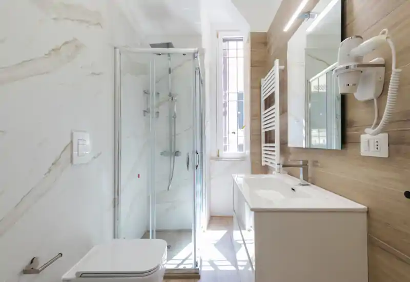 Salle de bains moderne et lumineuse avec cabine de douche vitrée, WC suspendu, meuble vasque avec miroir et sèche-cheveux ; carrelage effet marbre et panneaux imitation bois pour une ambiance contemporaine et fraîche.