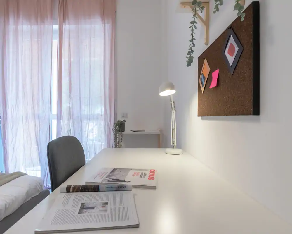 Bright, minimalist bedroom corner with a white desk, reading lamp and a neatly made bed partially visible; soft pink curtains diffuse natural light for a calm workspace.