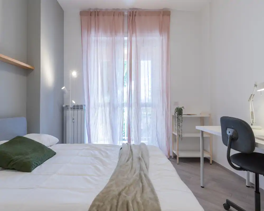 Bright, modern bedroom with a large bed dressed in white linens, soft throw and a green cushion; sheer pink curtains diffuse natural light and a tidy desk provides a work corner.