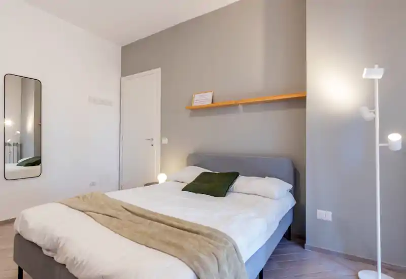 Bright, modern bedroom with a neatly made double bed, neutral gray accent wall and minimalist decor including a wooden shelf and floor lamp.
