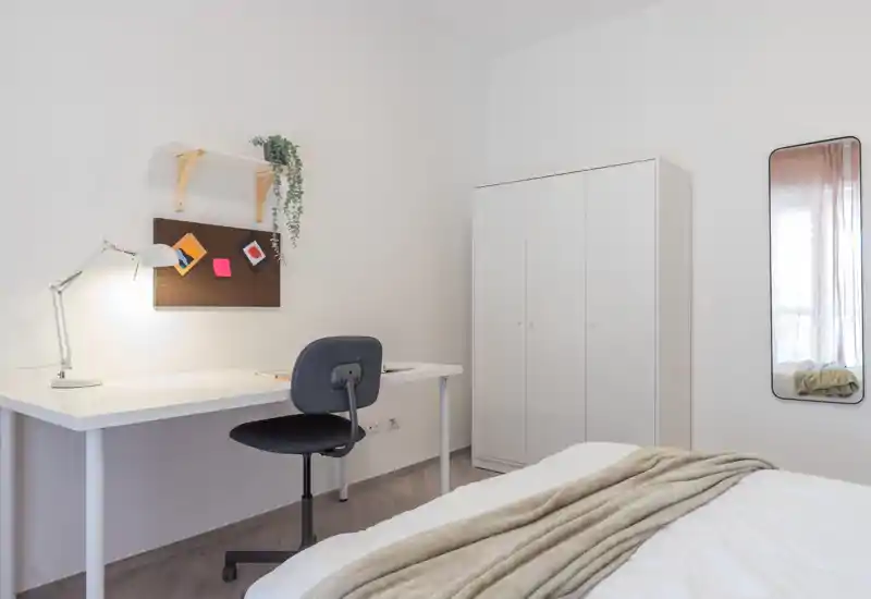 Minimalist bright bedroom with a neatly made bed in the foreground, a white wardrobe and a simple desk with reading lamp — ideal for a student or single occupant.