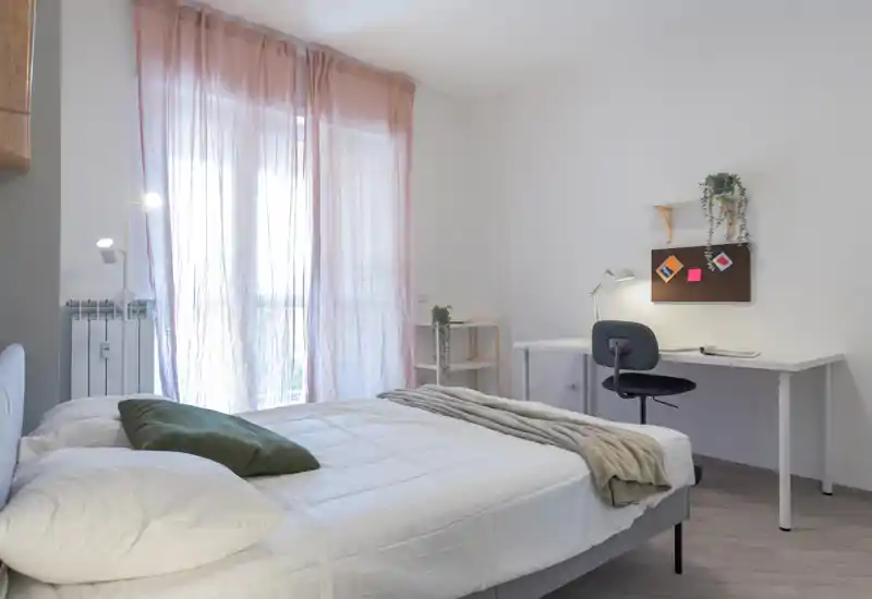 Bright, minimal bedroom featuring a neatly made double bed, soft natural light through sheer curtains, and a simple desk corner — ideal for rest and remote work.