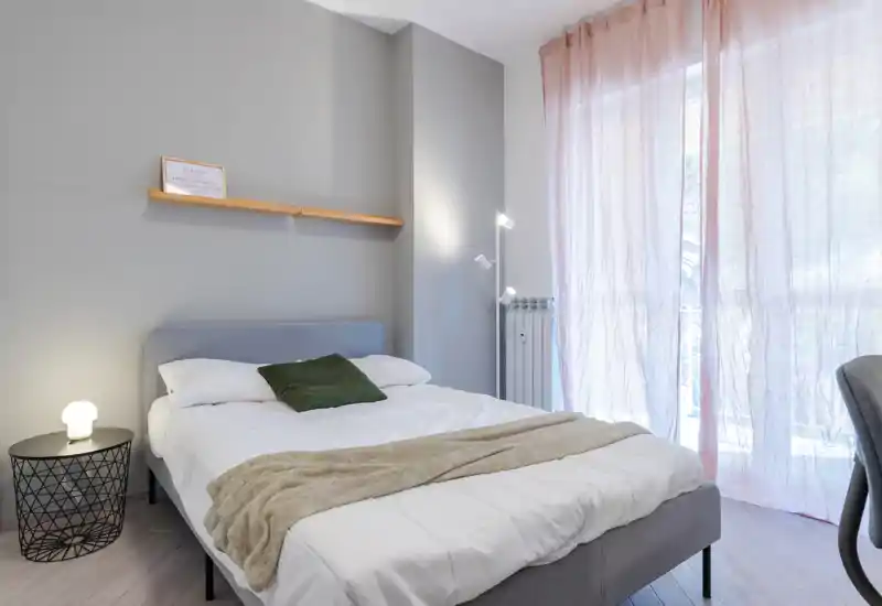 Bright, modern bedroom with a neatly made double bed, soft throw and cushion, bedside lamp and floor lamp; large window with sheer curtains provides abundant natural light.