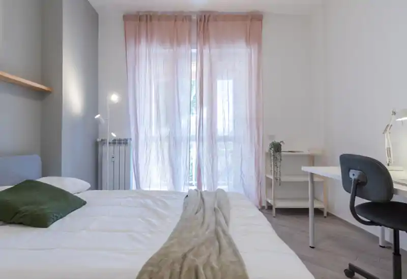 Bright, modern bedroom with a large bed dressed in white linens, soft throw and a green cushion; sheer pink curtains diffuse natural light and a tidy desk provides a work corner.