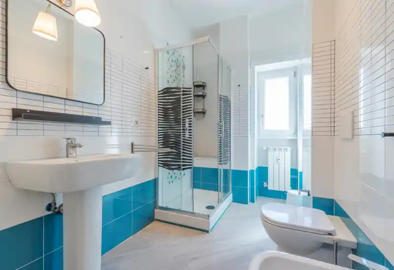 Salle de bains moderne et lumineuse avec cabine de douche en verre, toilettes murales, lavabo sur colonne et carrelage bleu et blanc ; lumière naturelle par la fenêtre.