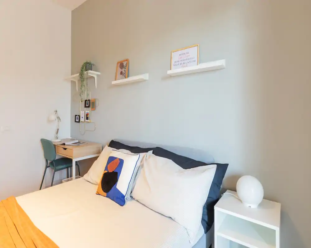 Dormitorio moderno y luminoso con cama doble bien hecha, cojines decorativos y una mesita de noche. Se aprecia un escritorio compacto y estantes que hacen la zona de descanso ordenada y funcional.