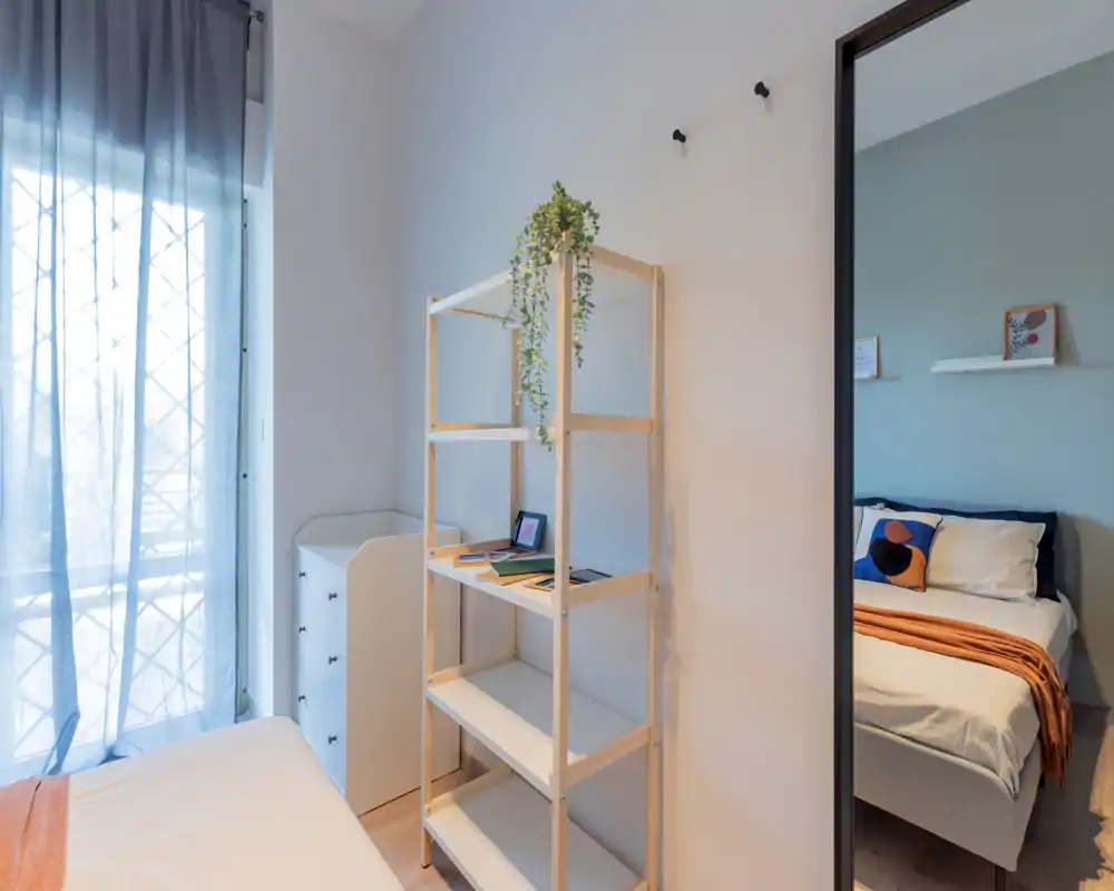 Dormitorio moderno y luminoso con luz natural y decoración cuidada: cama visible con cojines decorativos y manta cálida, estantería y ventana con cortinas traslúcidas que aportan sensación de amplitud y confort.