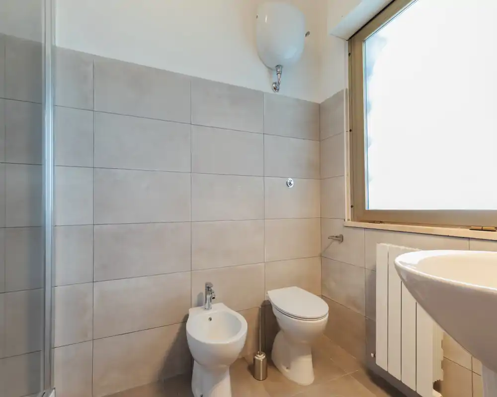 Bagno moderno e luminoso con wc, bidet, lavabo e una finestra opaca che lascia entrare luce naturale.