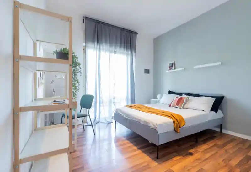 Camera da letto moderna e luminosa con letto matrimoniale in lenzuola bianche e un plaid senape, luce naturale da balcone e una scrivania minimalista.