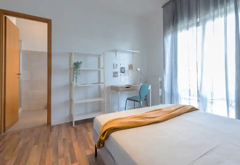 Camera da letto luminosa e moderna con letto ben rifatto, scrivania per lo studio e ampia finestra con tende leggere che lasciano entrare molta luce naturale.