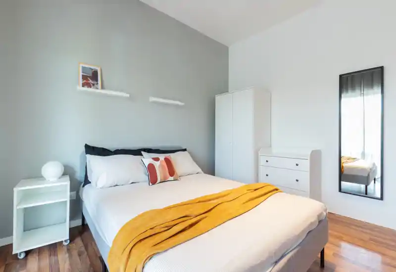 Camera da letto moderna e luminosa con letto matrimoniale ben rifatto, plaid colorato e cuscino decorativo, armadio e specchio a figura intera — perfetta per annunci di affitto.