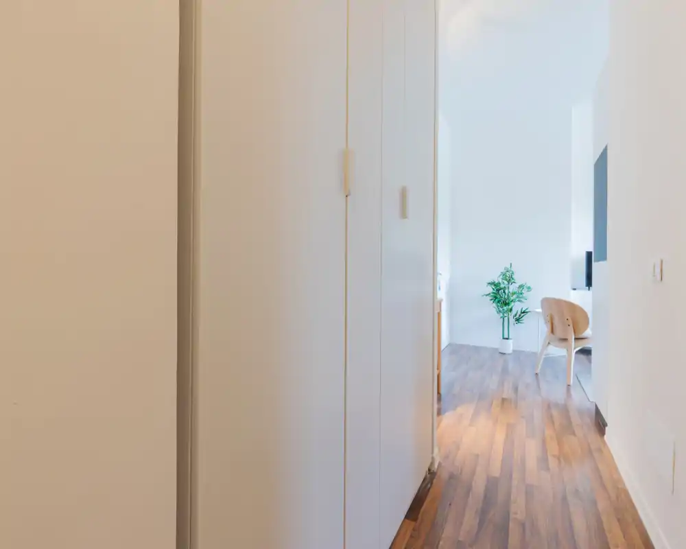Ingresso/corridoio luminoso e minimal con armadi a incasso e pavimento in legno che conduce a un'area giorno con una pianta e seduta visibili in fondo.