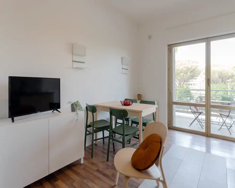 Zona giorno moderna e luminosa con tavolo in legno, sedie verdi, mobile TV e ampie porte scorrevoli che si aprono sul balcone — ben illuminata e arredata con cura per affitti.
