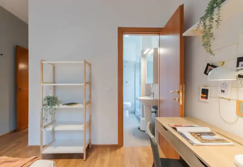 Rincón de dormitorio luminoso y compacto con escritorio de madera, estantería y baño en suite visible a través de la puerta abierta; estilo contemporáneo, limpio con plantas y tonos cálidos de madera.
