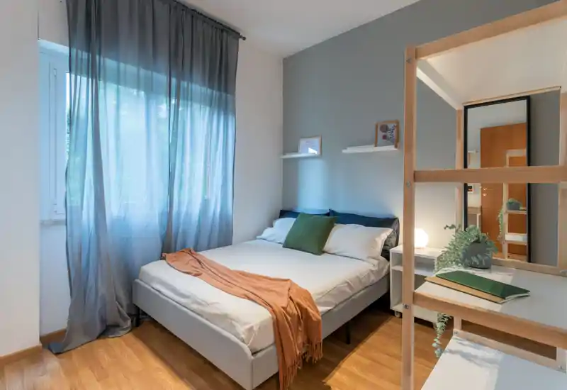 Dormitorio luminoso y bien decorado con una cama doble cómoda, textiles suaves y un escritorio/estantería junto a una gran ventana que aporta luz natural difusa.