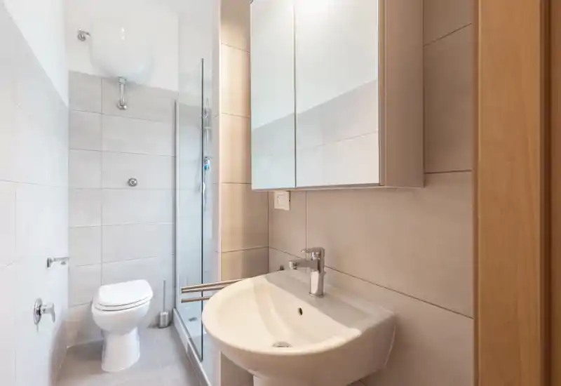 Baño moderno y compacto con inodoro suspendido, mampara de ducha de cristal y lavabo blanco bajo un armario con espejo — limpio, luminoso y bien iluminado.