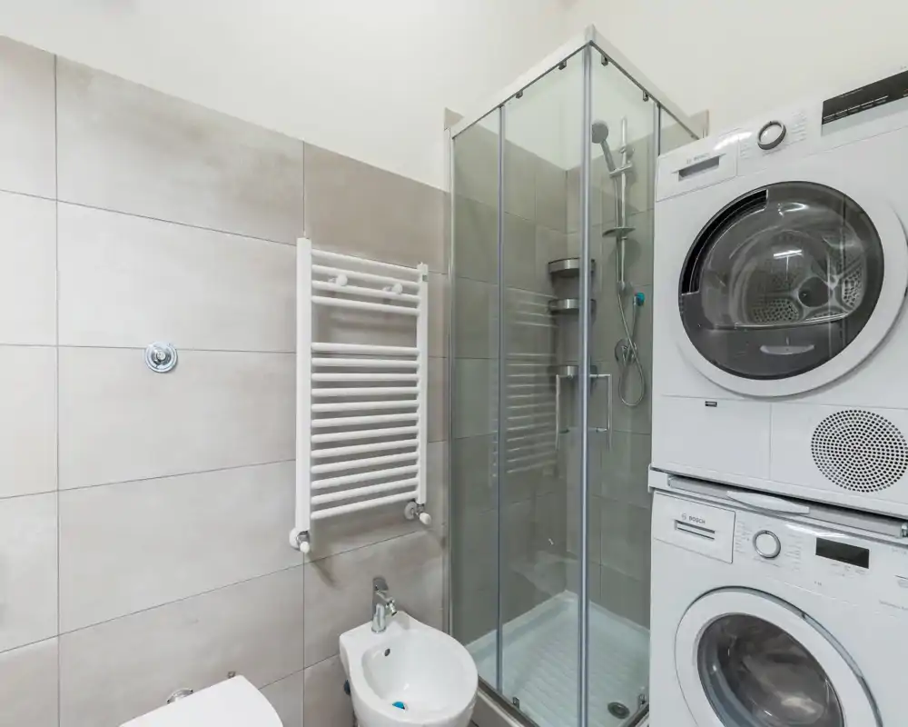 Compact modern bathroom with a glass-enclosed shower, wall-mounted towel radiator and stacked washer-dryer unit. Clean tiles and contemporary fixtures create a practical, well-maintained space.
