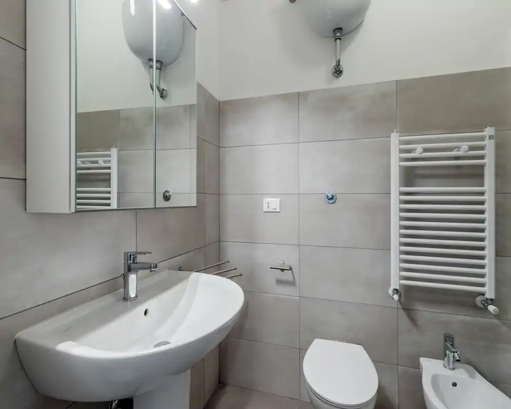 Modern, well-lit bathroom with a wall-mounted towel warmer, ceramic sink, toilet and bidet, and a mirrored cabinet above the sink.