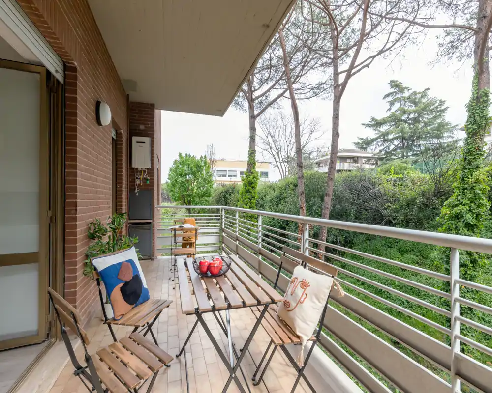 Sunny covered balcony with a wooden table and foldable chairs, decorated with cushions and a bowl of fruit; overlooks a green garden for relaxed outdoor dining.