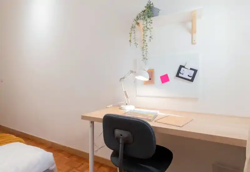 Compact bedroom corner with a neat wooden desk and lamp next to a partially visible bed; bright, minimal and functional workspace with a hanging shelf and plant.