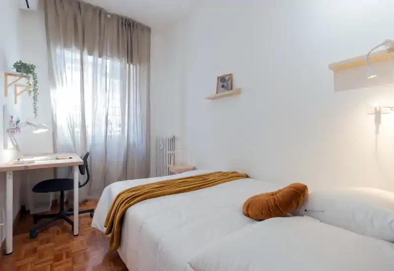 Bright, cozy single bedroom with a neatly made bed, warm throw and cushion, and a small work desk by the window letting in soft natural light.