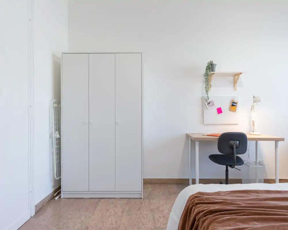 Rincón de dormitorio minimalista y bien iluminado con el borde de la cama visible, un armario blanco y un escritorio ordenado — luminoso y apropiado para anuncios.