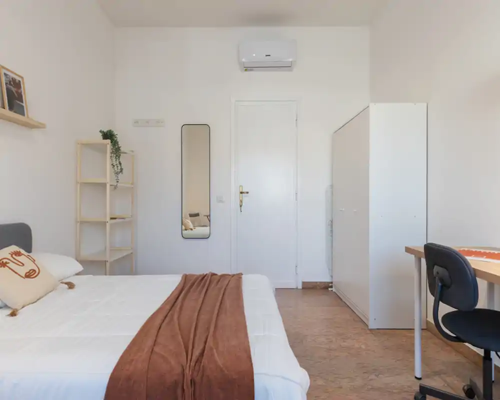 Dormitorio luminoso y minimalista con cama grande, manta cálida y almacenamiento sencillo; ideal para una estancia tranquila con zona de escritorio.
