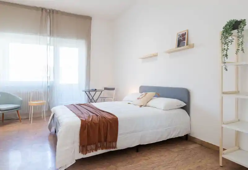 Dormitorio luminoso y minimalista con cama doble bien hecha, textiles suaves y luz natural de una gran ventana — acogedor y sereno.