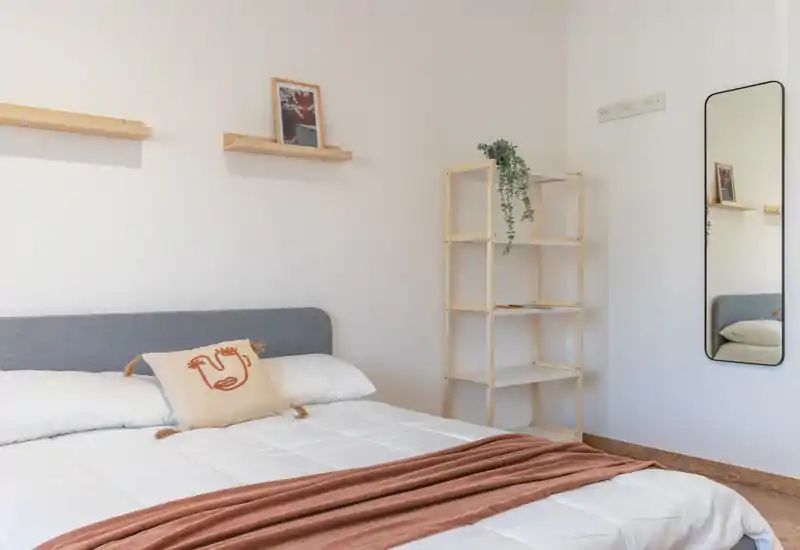 Dormitorio luminoso y minimalista con cama doble bien hecha, textiles neutros y una manta cálida. Estanterías de madera y un espejo alto completan el ambiente diáfano de estilo escandinavo.