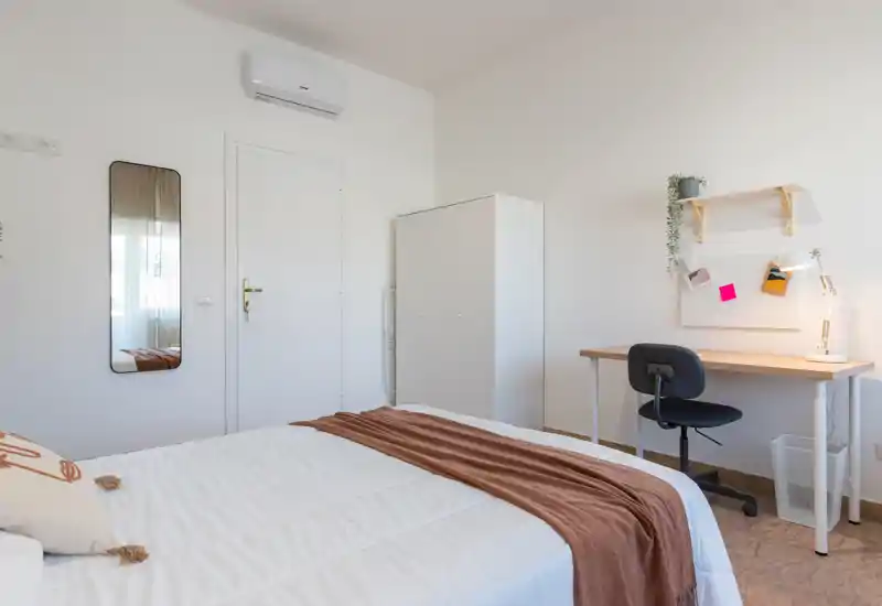 Dormitorio limpio y luminoso con cama doble, manta marrón, armario y un escritorio sencillo — ideal para una estancia cómoda o trabajo a distancia.