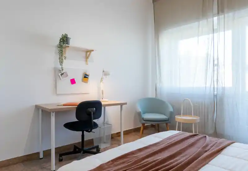 Dormitorio luminoso y minimalista con cama bien hecha, escritorio de trabajo y un sillón cómodo junto a una gran ventana que deja entrar luz natural suave.