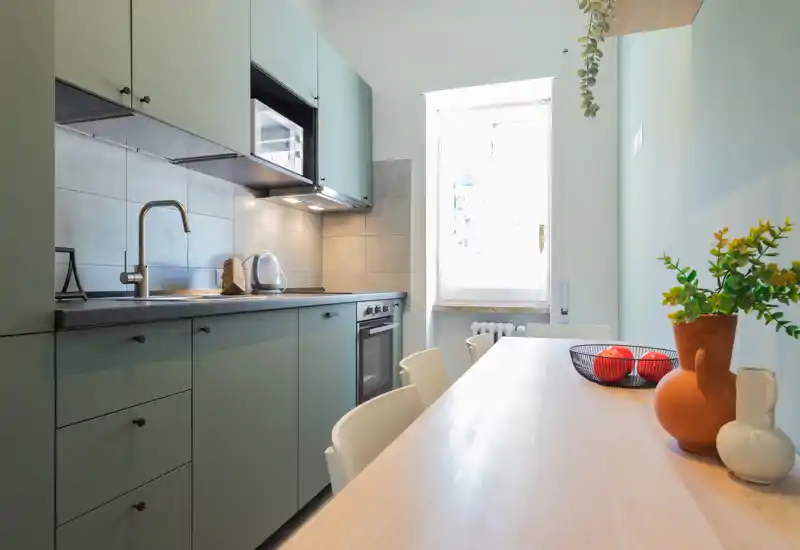 Bright, modern kitchen with sage-green cabinetry, integrated appliances and a wooden dining surface styled with a vase and fruit bowl — well lit by a window.
