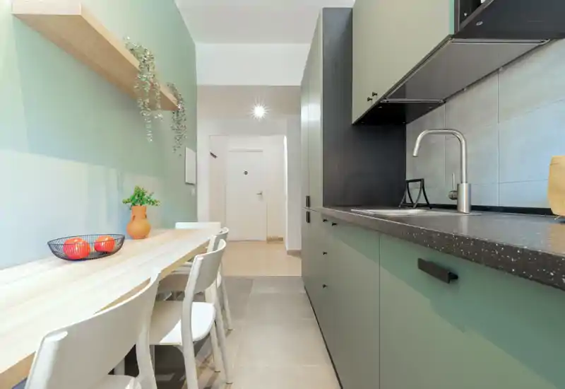 Modern galley-style kitchen with matte green cabinets, a dark stone countertop and an integrated sink. A light wood dining counter with chairs, potted plants and minimalist shelving create a fresh, welcoming dining nook.