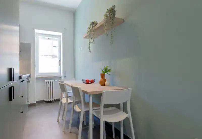 Compact dining corner with a light wood table, four white chairs and natural light from a window. Minimalist décor includes hanging plants and a small vase for a fresh, inviting breakfast nook.