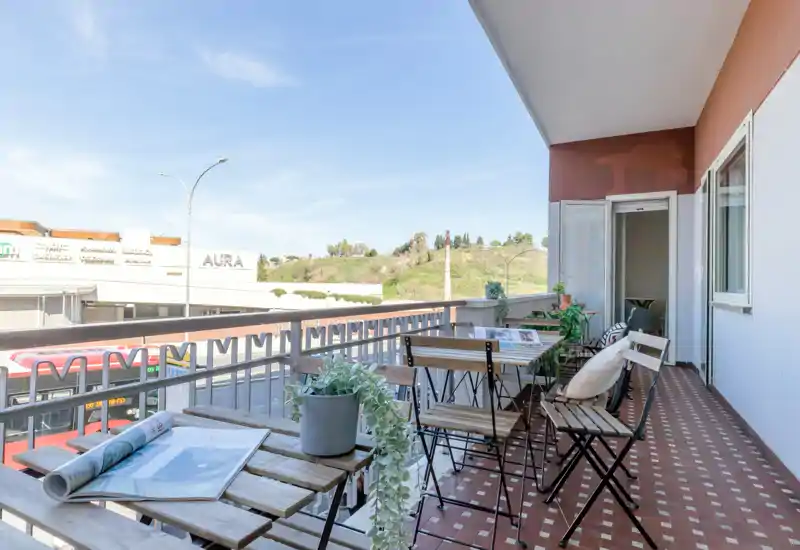Sunny furnished balcony with a table and folding chairs, potted plants and views over the street and nearby green hillside — ideal for breakfast or relaxing outdoors.