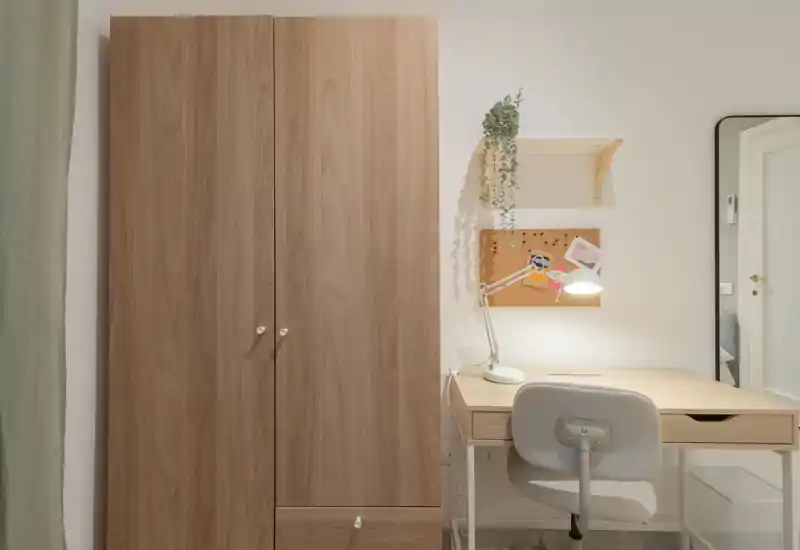 Rincón de dormitorio con un gran armario de madera y un escritorio compacto con lámpara y silla; luminoso, ordenado y suavemente decorado con planta colgante y corcho.